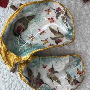 Peut inclure: Un plat décoratif en forme de coquille avec un bord doré. Le plat est décoré d'un fond blanc et d'un motif de colibris et de papillons dans des tons de vert, bleu et rouge.