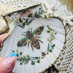 Puede incluir: Una concha de mar blanca con un borde dorado, decorada con una abeja colorida y diseños florales. La concha está en un soporte y se utiliza como plato decorativo.