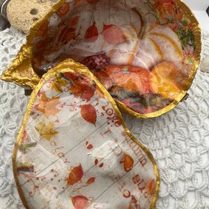 Może przedstawiać: Dwie ozdobne, pozłacane miski z papier-mache w jesiennym stylu. Miski są ozdobione białą dynią, pomarańczowymi i czerwonymi liśćmi oraz nadrukiem w stylu vintage. Miski stoją na białym, szydełkowym podkładzie.