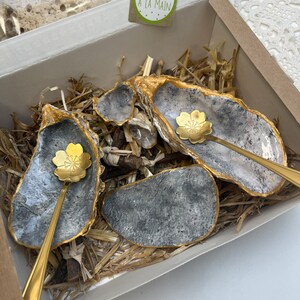 Puede incluir: Un juego de cuatro peque&ntilde;os platos de concha de ostra grises y dorados con cucharas doradas. Los platos est&aacute;n en una caja con material de embalaje de paja. Una peque&ntilde;a etiqueta con el texto "FAIT A LA MAIN" es visible en la esquina superior derecha de la imagen.