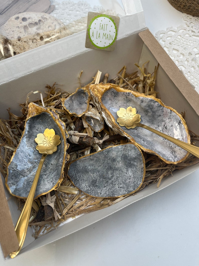 Puede incluir: Un juego de cuatro peque&ntilde;os platos de concha de ostra grises y dorados con cucharas doradas. Los platos est&aacute;n en una caja con material de embalaje de paja. Una peque&ntilde;a etiqueta con el texto "FAIT A LA MAIN" es visible en la esquina superior derecha de la imagen.