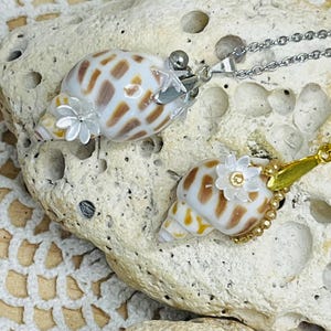 Peut inclure: Deux pendentifs coquillage avec chaînes en argent. Les coquillages sont blancs avec des marques marron et ornés de petits accents en forme de fleurs et d'étoiles blanches. Un pendentif a une chaîne en argent, l'autre une chaîne en or.