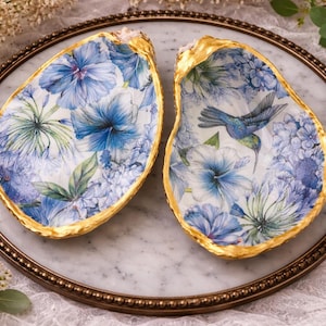 Peut inclure: Deux plats décoratifs en forme de coquilles d'huîtres avec un motif floral dans les tons de bleu et de vert, avec une bordure dorée. Une coquille représente un colibri. Les coquilles sont présentées sur un plateau en marbre.