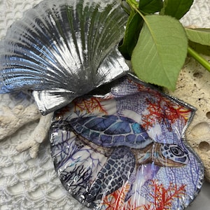 Puede incluir: Una concha de plata con un diseño detallado de una tortuga marina nadando en un arrecife de coral. La concha es una pieza decorativa con un acabado brillante.