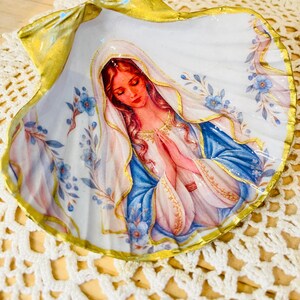 Puede incluir: Una concha de vieira decorativa con una imagen pintada de la Virgen Mar&iacute;a con las manos juntas en oraci&oacute;n. La figura lleva un velo blanco y una t&uacute;nica azul con adornos dorados. Los bordes de la concha est&aacute;n pintados en oro met&aacute;lico y la superficie est&aacute; decorada con peque&ntilde;as flores azules y hojas verdes sobre un fondo blanco.