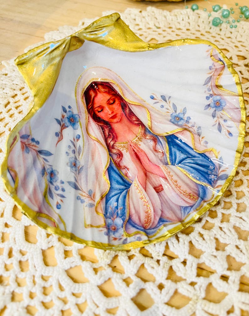 Puede incluir: Una concha de vieira decorativa con una imagen pintada de la Virgen Mar&iacute;a con las manos juntas en oraci&oacute;n. La figura lleva un velo blanco y una t&uacute;nica azul con adornos dorados. Los bordes de la concha est&aacute;n pintados en oro met&aacute;lico y la superficie est&aacute; decorada con peque&ntilde;as flores azules y hojas verdes sobre un fondo blanco.