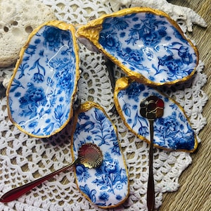 Op de afbeelding: Vier oesterschelpen met een blauw bloemenpatroon en gouden rand, gerangschikt op een wit kanten kleedje. Er zijn ook twee kleine, decoratieve lepels met bloemmotieven. De schelpen zijn een decoratief item.