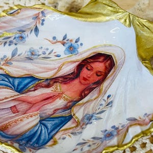Puede incluir: Una concha de ostra decorativa con una ilustraci&oacute;n pintada de una figura con cabello largo y un velo, con las manos juntas en oraci&oacute;n. La concha tiene un fondo blanco con detalles florales azules y un borde pintado en color oro met&aacute;lico.