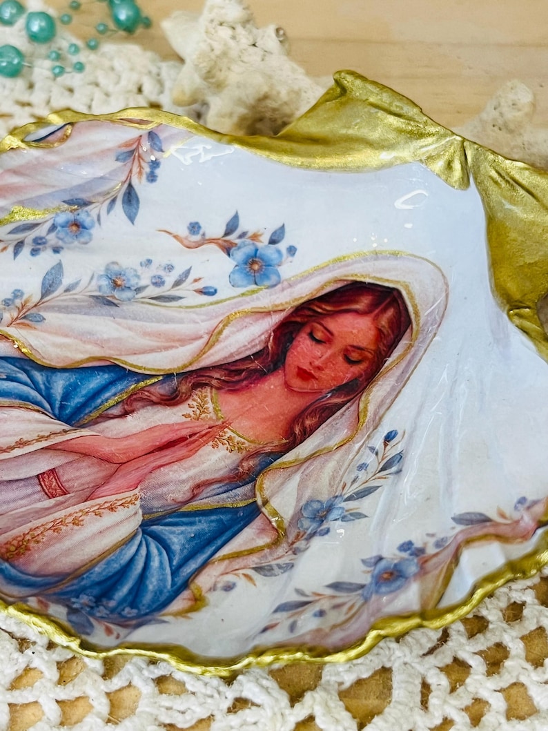Puede incluir: Una concha de ostra decorativa con una ilustraci&oacute;n pintada de una figura con cabello largo y un velo, con las manos juntas en oraci&oacute;n. La concha tiene un fondo blanco con detalles florales azules y un borde pintado en color oro met&aacute;lico.