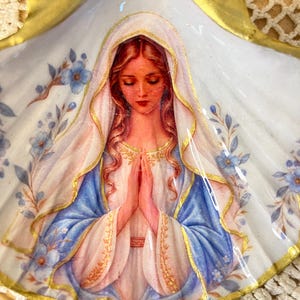 Puede incluir: Una concha de vieira decorativa con la imagen pintada de una figura de pelo largo, con velo blanco y t&uacute;nica azul, con las manos juntas en oraci&oacute;n. La concha tiene un borde pintado de dorado y est&aacute; decorada con peque&ntilde;as flores azules.