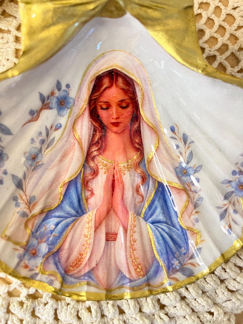 Puede incluir: Una concha de vieira decorativa con la imagen pintada de una figura de pelo largo, con velo blanco y t&uacute;nica azul, con las manos juntas en oraci&oacute;n. La concha tiene un borde pintado de dorado y est&aacute; decorada con peque&ntilde;as flores azules.