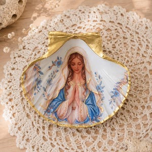 Puede incluir: Una concha de vieira decorativa con una ilustraci&oacute;n pintada de una figura de cabello casta&ntilde;o largo, con velo blanco y t&uacute;nica azul, con las manos juntas en oraci&oacute;n. El borde de la concha est&aacute; pintado de color dorado y tiene flores azules.