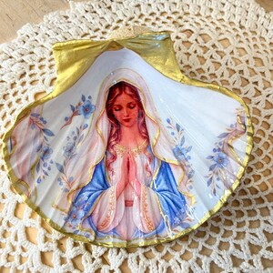 Puede incluir: Una concha marina decorativa con la imagen pintada de una figura de pelo largo que lleva un velo blanco y una capa azul, con las manos juntas en oraci&oacute;n. La concha tiene un borde pintado de dorado y peque&ntilde;as flores azules y hojas verdes.