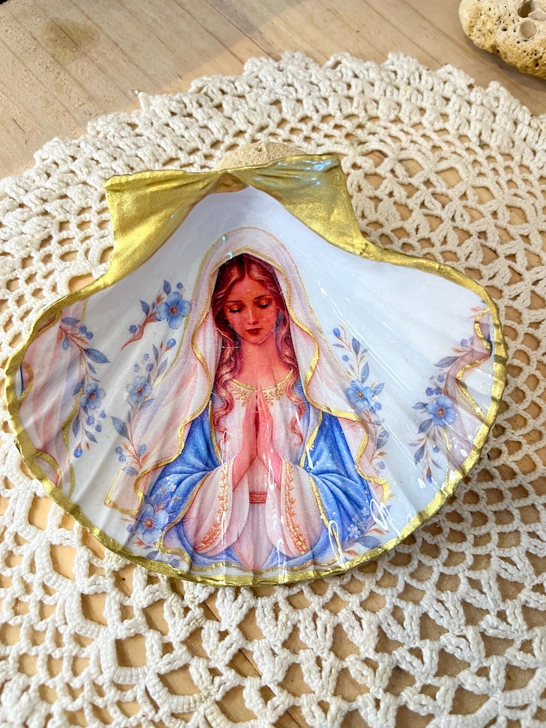 Puede incluir: Una concha marina decorativa con la imagen pintada de una figura de pelo largo que lleva un velo blanco y una capa azul, con las manos juntas en oraci&oacute;n. La concha tiene un borde pintado de dorado y peque&ntilde;as flores azules y hojas verdes.