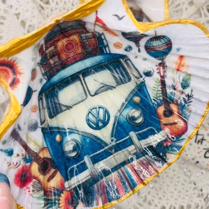 Op de afbeelding: Een witte schelp met een gouden rand, versierd met een blauwe en witte Volkswagen bus, een gitaar en een luchtballon. De bus heeft een VW-logo op de voorkant.