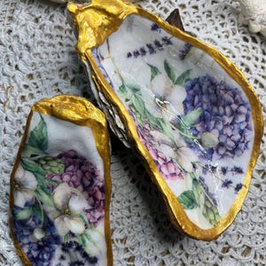Pode incluir: Duas conchas de ostra pintadas com uma borda dourada e um desenho floral com flores brancas, hortênsias roxas e rosadas e lavanda.