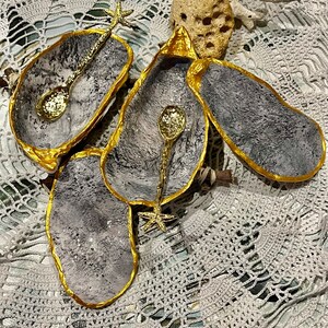 Puede incluir: Tres cuencos de concha de ostra grises y dorados con asas de estrella de mar doradas y cucharas doradas. Los cuencos est&aacute;n sobre una superficie de crochet blanco.