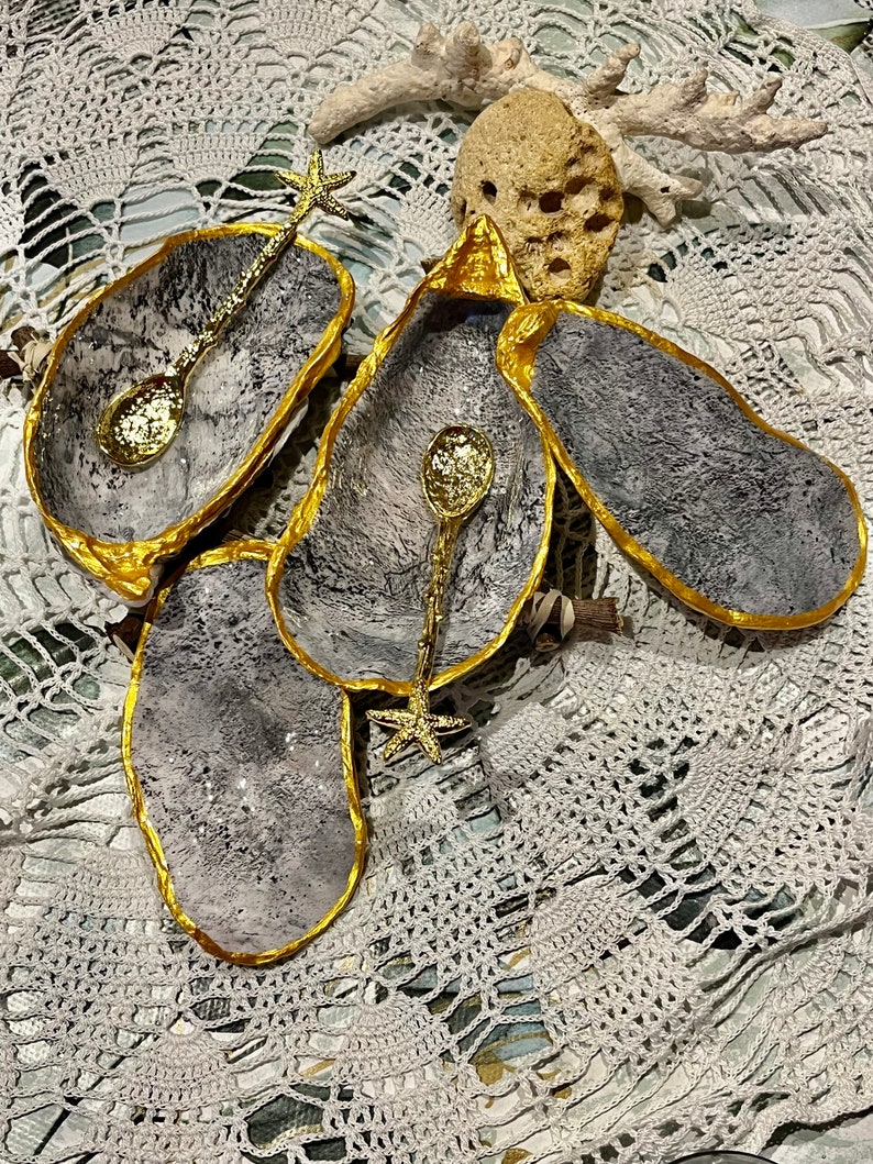 Puede incluir: Tres cuencos de concha de ostra grises y dorados con asas de estrella de mar doradas y cucharas doradas. Los cuencos est&aacute;n sobre una superficie de crochet blanco.