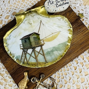 May include: A decorative seashell with a gold-painted rim, featuring a watercolour painting of a coastal structure. The shell rests on a wooden board, accompanied by a pebble with text and nautical-themed metal embellishments.