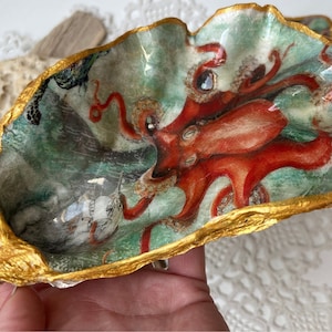 Può includere: Un piatto a forma di conchiglia di ostrica con un bordo dorato e un'immagine dipinta di un polpo in una scena di mare. Il polpo è rosso e ha tentacoli.