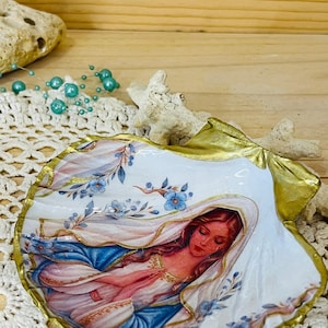 Puede incluir: Una concha marina decorativa con una ilustraci&oacute;n pintada de una figura de pelo largo con velo blanco y t&uacute;nica azul, con las manos juntas en oraci&oacute;n. La concha tiene un borde festoneado pintado en oro y detalles florales azules.
