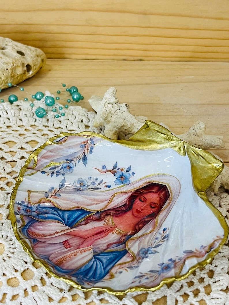 Puede incluir: Una concha marina decorativa con una ilustraci&oacute;n pintada de una figura de pelo largo con velo blanco y t&uacute;nica azul, con las manos juntas en oraci&oacute;n. La concha tiene un borde festoneado pintado en oro y detalles florales azules.
