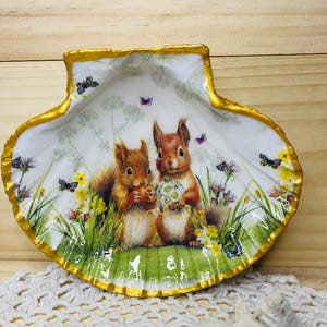 May include: A white seashell dish with a gold rim, featuring a painted scene of two squirrels in a green meadow with flowers and butterflies.