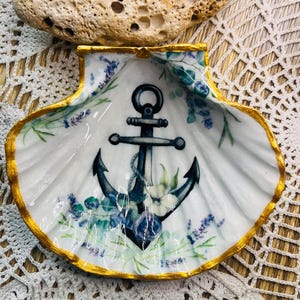 May include: A white seashell dish with a gold rim and a blue anchor design. The anchor is surrounded by floral details in shades of blue, green, and purple.