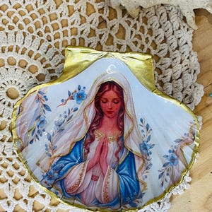 Puede incluir: Una concha de vieira decorativa con una ilustraci&oacute;n detallada de una figura de pelo largo con velo blanco y t&uacute;nica azul, con las manos juntas en oraci&oacute;n. La concha est&aacute; decorada con motivos florales azules y un borde pintado en oro met&aacute;lico.