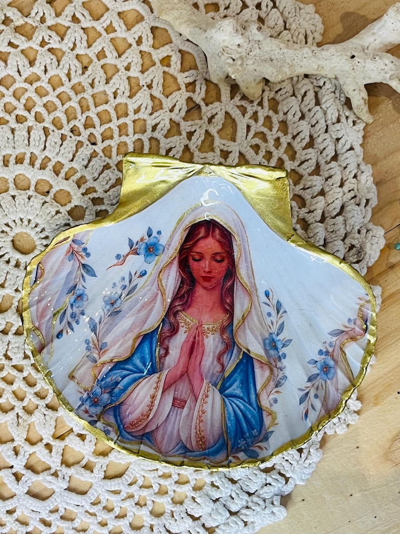 Puede incluir: Una concha de vieira decorativa con una ilustraci&oacute;n detallada de una figura de pelo largo con velo blanco y t&uacute;nica azul, con las manos juntas en oraci&oacute;n. La concha est&aacute; decorada con motivos florales azules y un borde pintado en oro met&aacute;lico.
