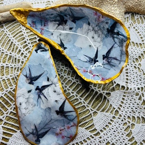 Könnte beinhalten: Zwei dekorative Austernmuscheln mit einem blauen und weißen Blumenmuster und goldenen Akzenten. Die Muscheln sind geöffnet und zeigen ein Design von Vögeln im Flug.