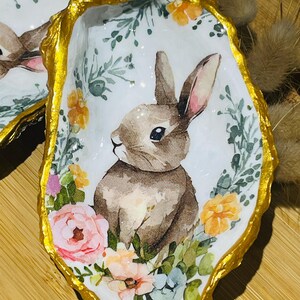 Può includere: Un guscio di ostrica in ceramica bianca con un bordo dorato, che presenta un dipinto ad acquerello di un coniglio marrone circondato da fiori rosa, gialli e blu.