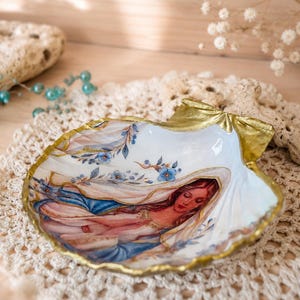 Puede incluir: Un plato de concha decorativo con una ilustraci&oacute;n pintada de una figura de pelo largo con un velo azul y blanco, decorado con peque&ntilde;as flores azules. Los bordes de la concha est&aacute;n pintados con un acabado en color oro met&aacute;lico.