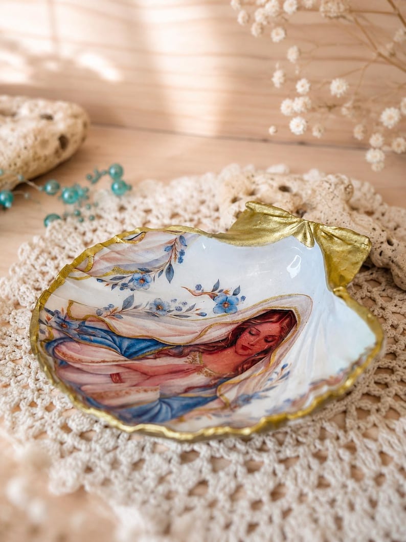 Puede incluir: Un plato de concha decorativo con una ilustraci&oacute;n pintada de una figura de pelo largo con un velo azul y blanco, decorado con peque&ntilde;as flores azules. Los bordes de la concha est&aacute;n pintados con un acabado en color oro met&aacute;lico.
