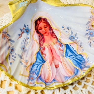 Puede incluir: Una concha de vieira decorativa con una ilustraci&oacute;n central de una figura con cabello largo y velo, con las manos juntas en oraci&oacute;n. La figura viste una prenda blanca y azul. Los bordes de la concha est&aacute;n pintados de dorado con detalles florales.