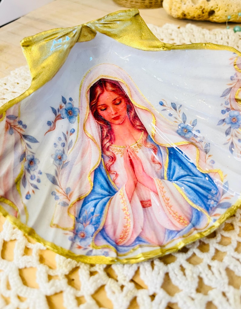 Puede incluir: Una concha de vieira decorativa con una ilustraci&oacute;n central de una figura con cabello largo y velo, con las manos juntas en oraci&oacute;n. La figura viste una prenda blanca y azul. Los bordes de la concha est&aacute;n pintados de dorado con detalles florales.