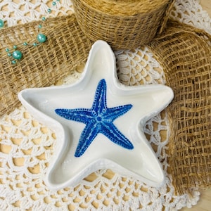 Può includere: Un piatto a forma di stella marina in ceramica bianca con un disegno di stella marina blu al centro. Il piatto è su un centrino all'uncinetto bianco, con un nastro di juta e una bobina di spago sullo sfondo. Sono presenti anche perline decorative.