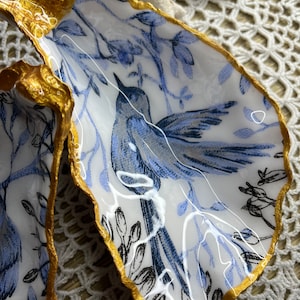 Puede incluir: Dos conchas de ostra blancas con ribete dorado, cada una con un diseño floral azul y blanco con un pájaro en el centro.