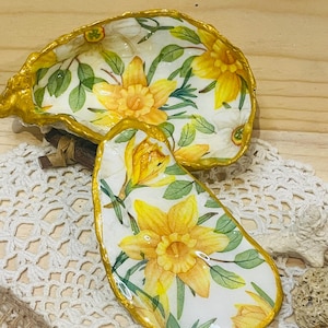 May include: Two decorative oyster shell dishes. The shells are painted with yellow daffodils and green leaves on a white background. The edges are trimmed with gold paint. One shell is oval, and the other is a half-shell shape.