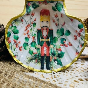Peut inclure: Un plat décoratif en coquillage avec un motif de soldat casse-noisette peint. L'intérieur est blanc avec une illustration festive du casse-noisette, de baies rouges et de feuillage vert. Le bord de la coquille est orné de peinture dorée, créant un accent sur le thème des fêtes.