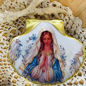 Puede incluir: Una concha de vieira con una imagen de decoupage de una mujer de pelo largo con velo blanco y t&uacute;nica azul, con las manos juntas en oraci&oacute;n. La concha est&aacute; decorada con motivos florales azules y tiene un borde pintado en oro met&aacute;lico.