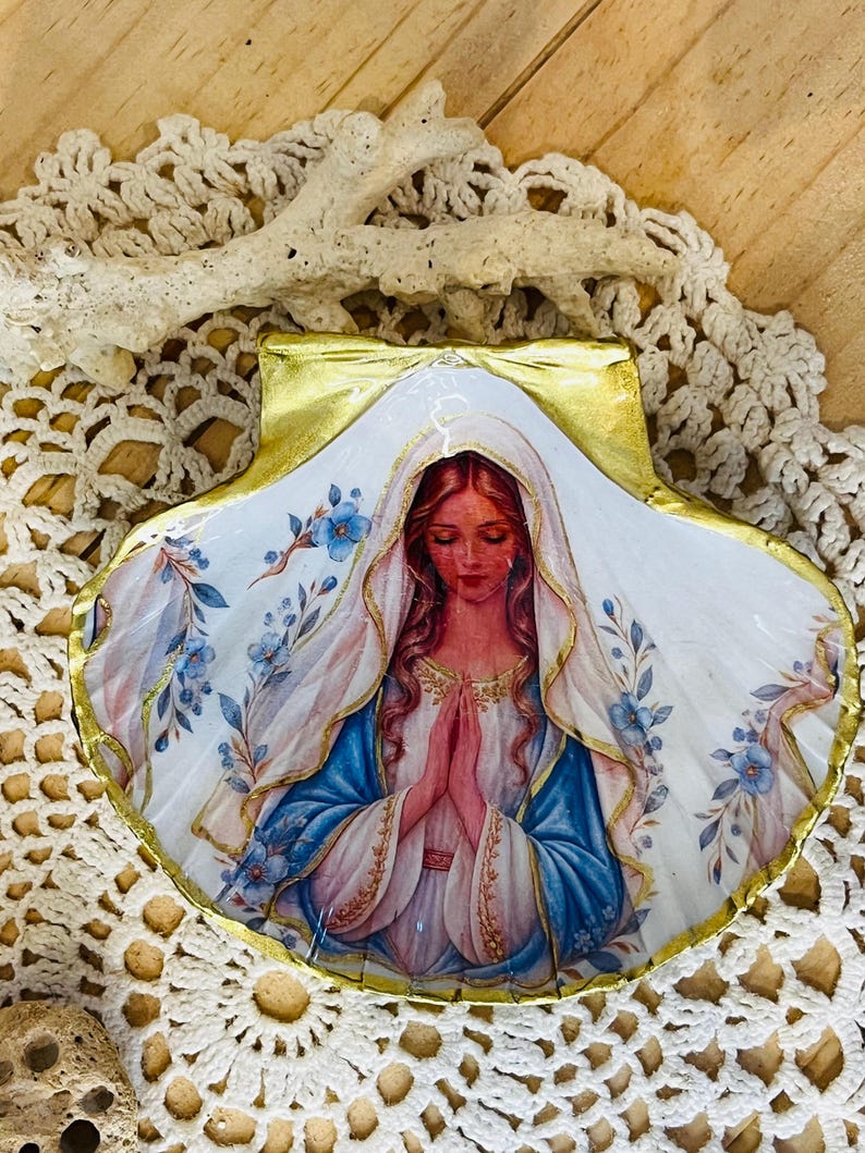 Puede incluir: Una concha de vieira con una imagen de decoupage de una mujer de pelo largo con velo blanco y t&uacute;nica azul, con las manos juntas en oraci&oacute;n. La concha est&aacute; decorada con motivos florales azules y tiene un borde pintado en oro met&aacute;lico.