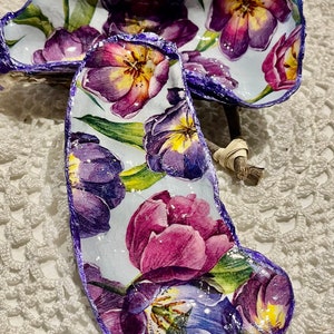 May include: Two decorative, purple-rimmed, white porcelain shells with a floral design of purple, pink, and yellow tulips. The shells are on a white crocheted surface.