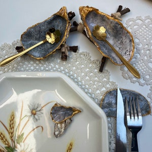 Puede incluir: Dos conchas de ostra con borde dorado y un interior gris, cada una con una peque&ntilde;a cuchara dorada en su interior. Las conchas est&aacute;n montadas en peque&ntilde;as ramas de madera. Una tercera concha de ostra est&aacute; en un plato blanco con un dise&ntilde;o floral. Un tenedor y un cuchillo est&aacute;n en una concha de ostra gris.