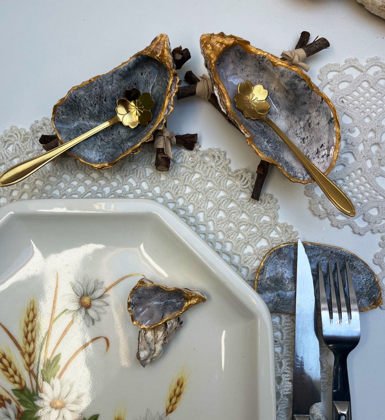 Puede incluir: Dos conchas de ostra con borde dorado y un interior gris, cada una con una peque&ntilde;a cuchara dorada en su interior. Las conchas est&aacute;n montadas en peque&ntilde;as ramas de madera. Una tercera concha de ostra est&aacute; en un plato blanco con un dise&ntilde;o floral. Un tenedor y un cuchillo est&aacute;n en una concha de ostra gris.