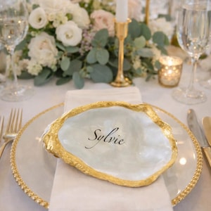 Può includere: Un'apparecchiatura con un tovagliolo bianco, un piatto trasparente con bordo dorato e una conchiglia decorativa con il nome "Sylvie". Il guscio ha un bordo dorato. Un'arrangiamento floreale e candele sullo sfondo.