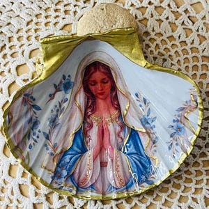 Puede incluir: Una concha marina decorativa con una imagen de decoupage de una figura de pelo largo, vestida con una t&uacute;nica blanca y azul con velo, con las manos juntas en oraci&oacute;n. La concha tiene un borde pintado en oro y detalles de flores azules.
