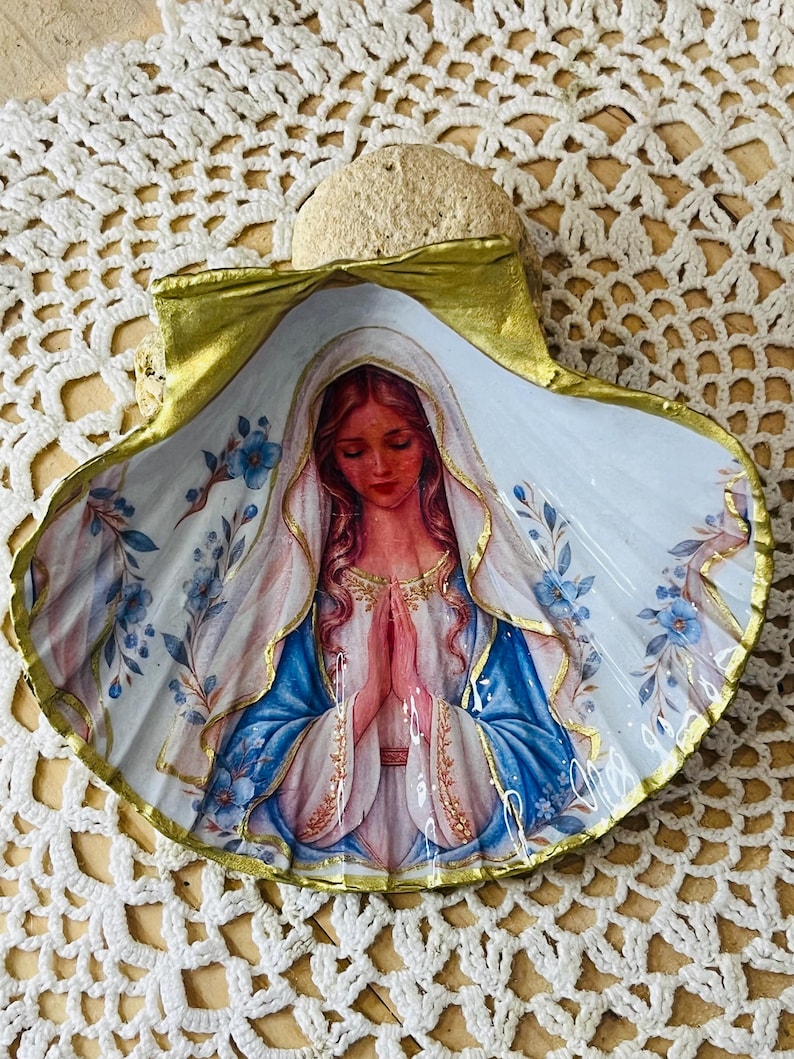 Puede incluir: Una concha marina decorativa con una imagen de decoupage de una figura de pelo largo, vestida con una t&uacute;nica blanca y azul con velo, con las manos juntas en oraci&oacute;n. La concha tiene un borde pintado en oro y detalles de flores azules.