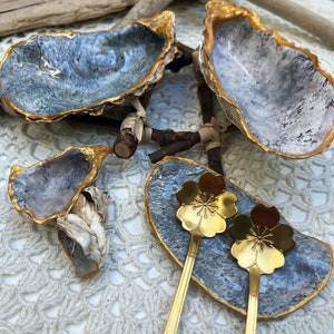 Puede incluir: Dos platos de concha de ostra con bordes dorados y un interior gris y blanco. Un plato est&aacute; lleno de dos cucharas doradas con un dise&ntilde;o de flor de cerezo. Los platos est&aacute;n sobre una superficie de crochet blanco.