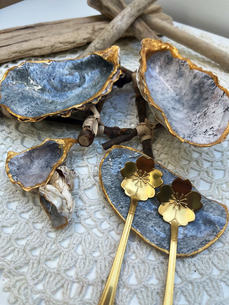Puede incluir: Dos platos de concha de ostra con bordes dorados y un interior gris y blanco. Un plato est&aacute; lleno de dos cucharas doradas con un dise&ntilde;o de flor de cerezo. Los platos est&aacute;n sobre una superficie de crochet blanco.